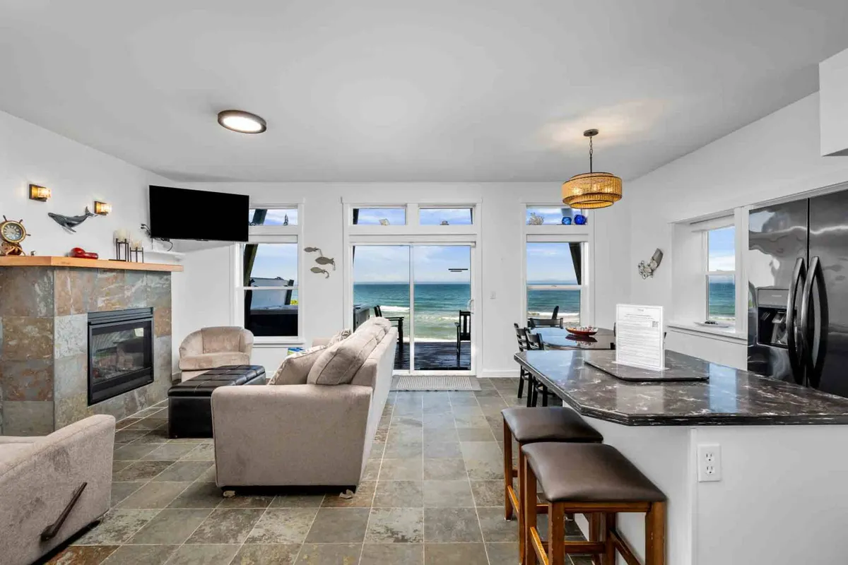 This interior hero image showcases a bright, open living space with expansive windows framing an uninterrupted view of the Pacific Ocean. The modern kitchen island invites casual dining, while a cozy fireplace anchors the room, creating an inviting space for relaxation. Sunlight pours in, illuminating the stylish furnishings and highlighting the property's seamless blend of comfort and coastal beauty.