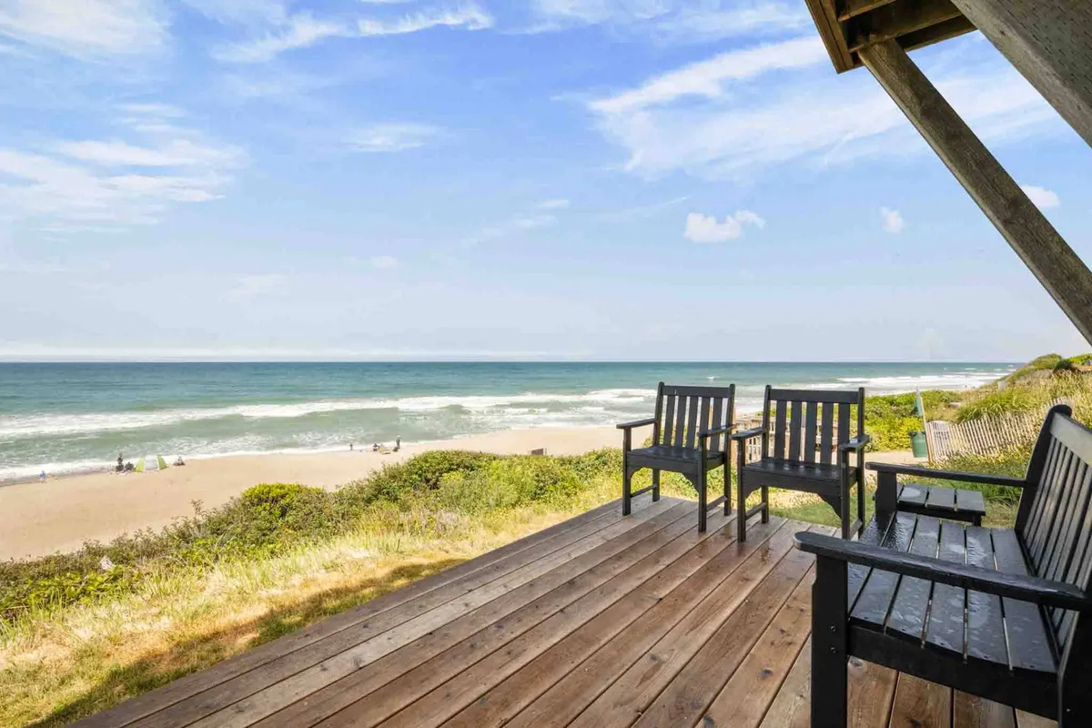 This exterior image features a sun-washed deck with comfortable seating overlooking the sand and ocean, with the beach just steps away. The scene captures the essence of easy beach access and the inviting outdoor environment, perfect for families and friends to relax after a day of adventures.