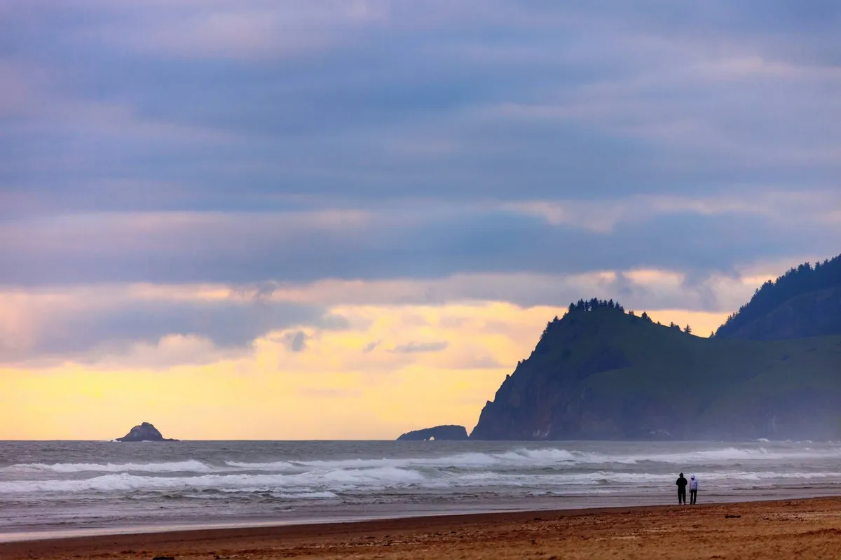 A romantic scene featuring a couple walking hand-in-hand along a vast sandy beach, with distant cliffs and a rocky island in the water under a cloudy sky. The shoreline stretches into the distance, offering a feeling of intimacy and natural beauty.
