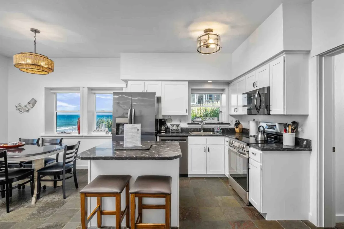 A cheerful kitchen and dining area featuring an island countertop, modern appliances, a spacious dining table, and windows offering glimpses of the sea. The space is designed for both cooking and socializing.