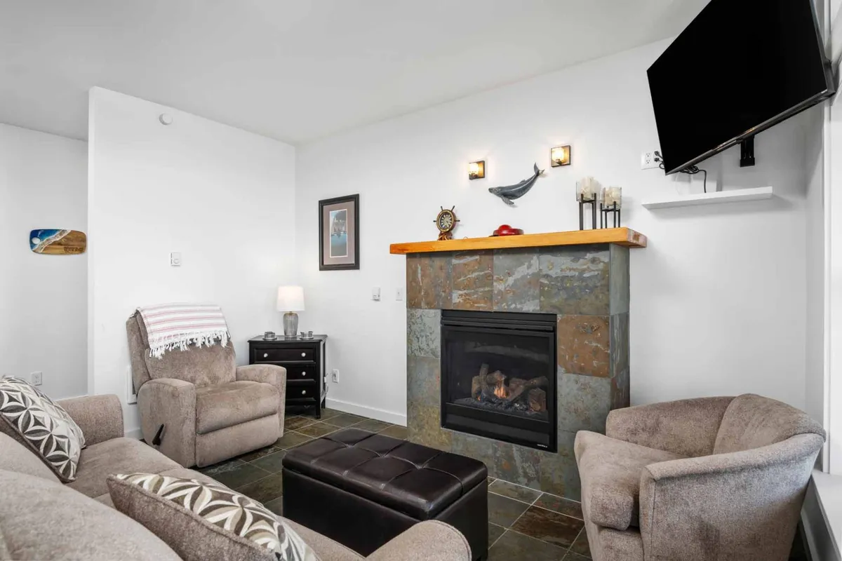 A warm, welcoming living room with a plush seating arrangement around a fireplace, a Smart TV, and decorative art. The space is bathed in natural light from white-trimmed windows, creating a cozy spot for relaxation or social gatherings.
