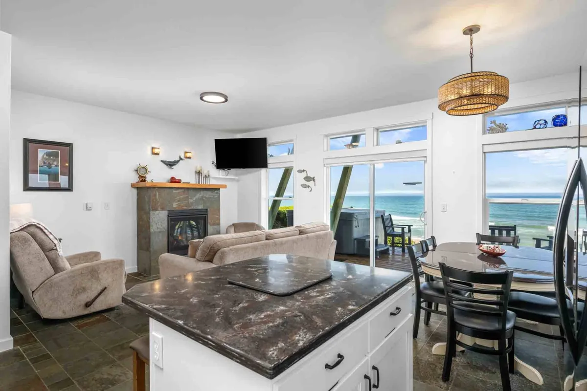 An inviting, open-concept living and dining area with a fireplace, large windows framing the ocean, and a dining table suited for group gatherings. The space feels cozy yet spacious, ideal for post-beach relaxation or shared meals.