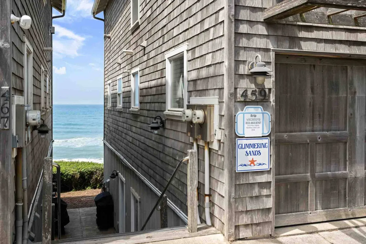 A tranquil exterior featuring the weathered wooden siding of the Glimmering Sands property, an ocean view, and a welcoming sign. The image evokes the sense of a secluded, exclusive community steps from the sand.