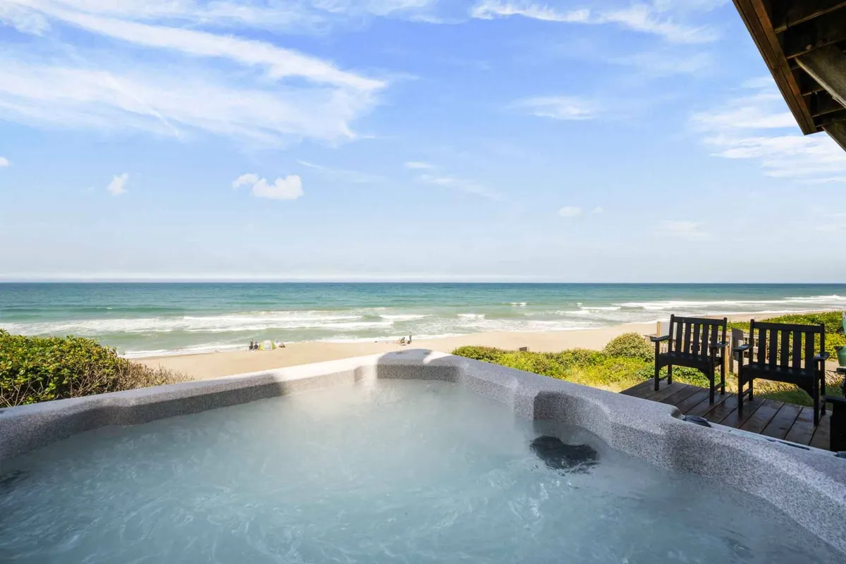 This image captures a serene oceanfront deck featuring a large private hot tub with sweeping views of the Pacific Ocean. The seating area is positioned to take in the sight of the waves, and the tranquil mood is set by golden sunlight, ideal for both relaxing in the hot tub and unwinding with a book or a coffee.