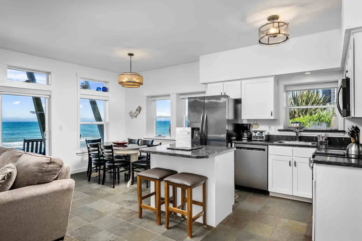 An inviting open-plan space where the kitchen island, dining table, and comfortable living room sofa are oriented towards large windows with sweeping ocean views, bringing the tranquility of the Pacific indoors.