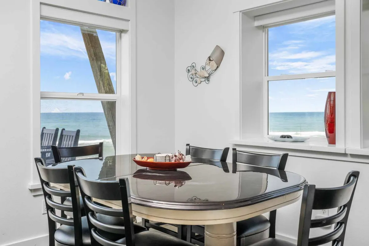 A sunlit dining table sits beside large windows that overlook the ocean, with decorative wall art and a vibrant fruit centerpiece creating a cheerful, welcoming mood. The setting is perfect for shared meals, celebrations, or simply relaxing together while enjoying views of the nearby beach.